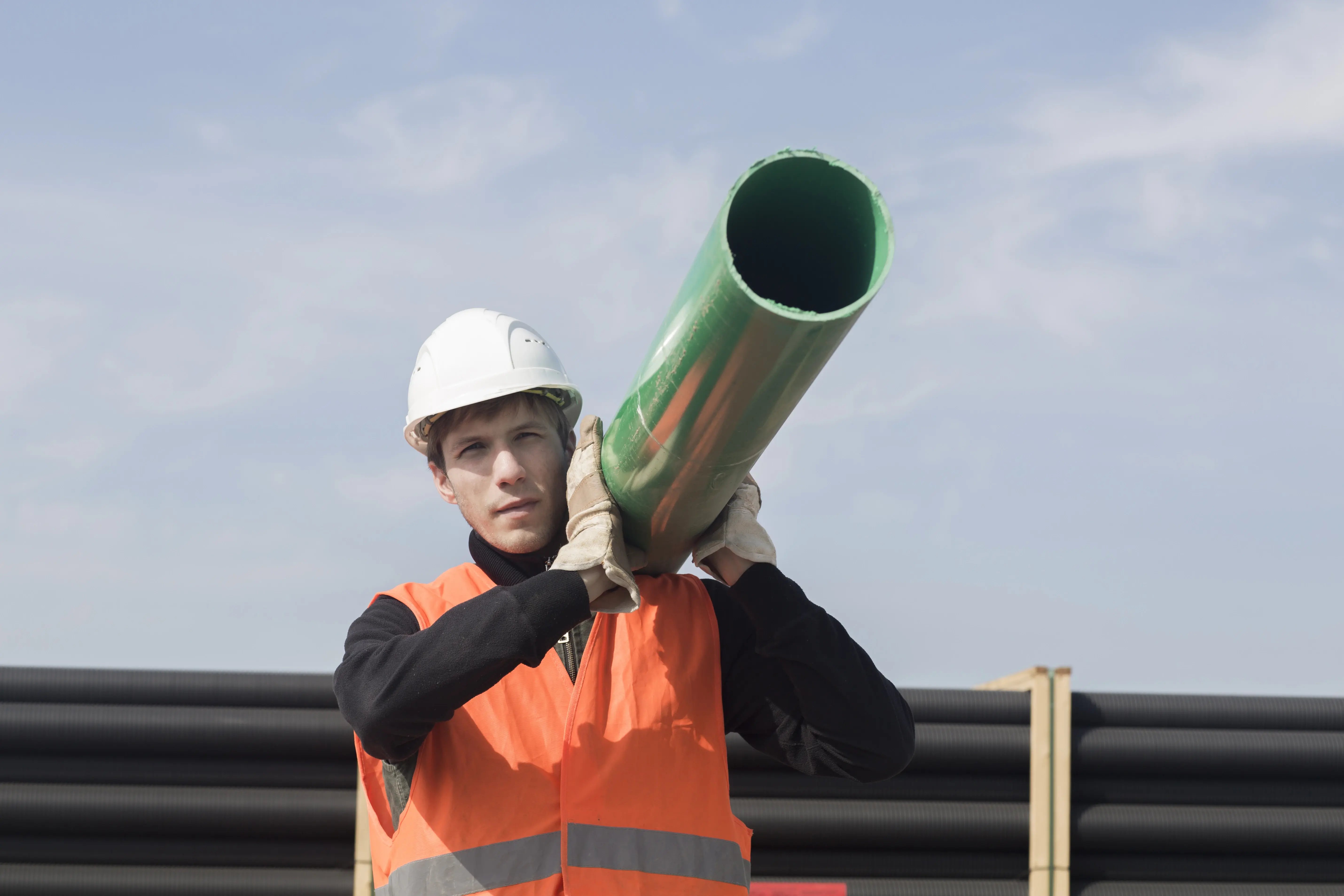 Construction worker wearing a safety vest and hard hat carrying a green pipeline section on his shoulder